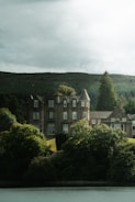 A large, old-fashioned stone mansion is surrounded by thick, lush greenery with a backdrop of rolling hills under a cloudy sky. The mansion features multiple windows and a prominent tower, exuding a sense of historical charm.
