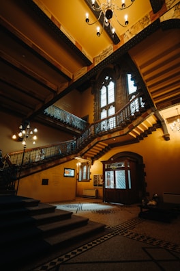 An elegant interior featuring a grand, ornate staircase within a historic building, complete with metal railings and decorative chandeliers. The room is lit with warm lighting which highlights the architectural details, including large arched windows and intricate moldings.