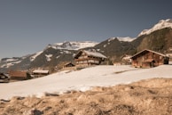 Several wooden cabins are situated on a snow-covered hillside, with majestic mountains in the background. The sky is clear and blue, creating a serene and picturesque alpine setting.