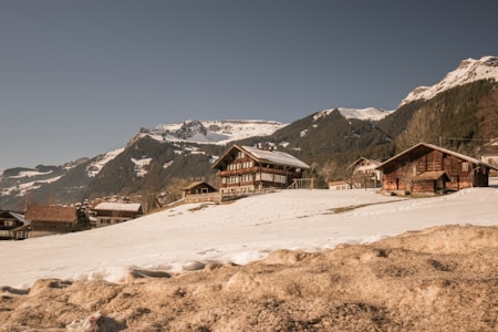 Several wooden cabins are situated on a snow-covered hillside, with majestic mountains in the background. The sky is clear and blue, creating a serene and picturesque alpine setting.
