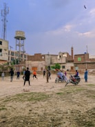 A group of people are playing cricket on an open dusty ground surrounded by urban buildings. Some children and adults are watching or participating in the game. There are motorcycles parked near the play area, and the backdrop includes a water tank and a cell tower. The sky is partly cloudy with a bird flying above.
