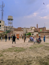 A group of people are playing cricket on an open dusty ground surrounded by urban buildings. Some children and adults are watching or participating in the game. There are motorcycles parked near the play area, and the backdrop includes a water tank and a cell tower. The sky is partly cloudy with a bird flying above.