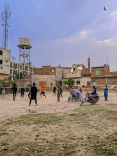 A group of people are playing cricket on an open dusty ground surrounded by urban buildings. Some children and adults are watching or participating in the game. There are motorcycles parked near the play area, and the backdrop includes a water tank and a cell tower. The sky is partly cloudy with a bird flying above.