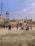 A group of people are playing cricket on an open dusty ground surrounded by urban buildings. Some children and adults are watching or participating in the game. There are motorcycles parked near the play area, and the backdrop includes a water tank and a cell tower. The sky is partly cloudy with a bird flying above.