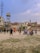 A group of people are playing cricket on an open dusty ground surrounded by urban buildings. Some children and adults are watching or participating in the game. There are motorcycles parked near the play area, and the backdrop includes a water tank and a cell tower. The sky is partly cloudy with a bird flying above.
