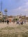 A group of people are playing cricket on an open dusty ground surrounded by urban buildings. Some children and adults are watching or participating in the game. There are motorcycles parked near the play area, and the backdrop includes a water tank and a cell tower. The sky is partly cloudy with a bird flying above.