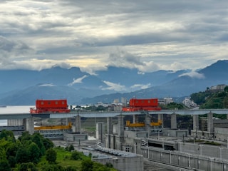 A massive dam structure with red machinery components sits amidst lush greenery and a backdrop of layered mountains and a cloudy sky. Modern buildings are visible in the distance, suggesting a blend of nature and industrial development.