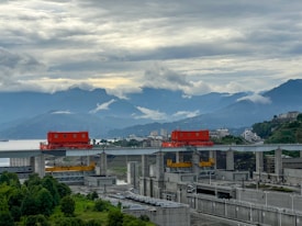 A massive dam structure with red machinery components sits amidst lush greenery and a backdrop of layered mountains and a cloudy sky. Modern buildings are visible in the distance, suggesting a blend of nature and industrial development.
