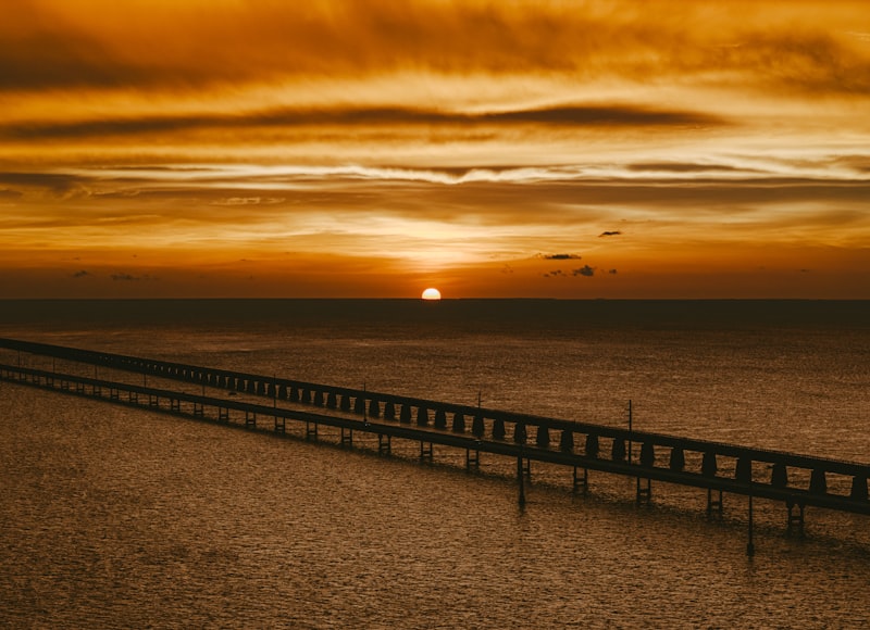 Silhouette of a long low bridge stretching across calm water beneath a deep orange sunset sky, the sun touching the horizon