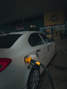 A car is parked at a gas station with a yellow fuel nozzle inserted into its tank. The setting is dimly lit and the background features signage from a store or restaurant.