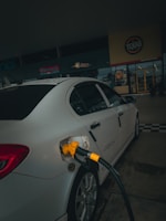 A car is parked at a gas station with a yellow fuel nozzle inserted into its tank. The setting is dimly lit and the background features signage from a store or restaurant.