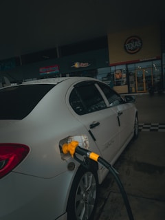 A car is parked at a gas station with a yellow fuel nozzle inserted into its tank. The setting is dimly lit and the background features signage from a store or restaurant.