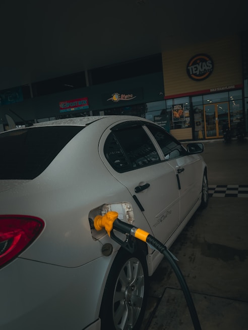 A car is parked at a gas station with a yellow fuel nozzle inserted into its tank. The setting is dimly lit and the background features signage from a store or restaurant.