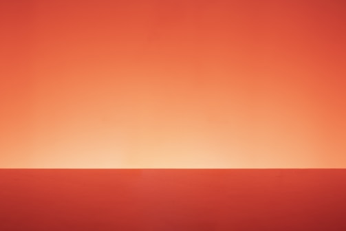 A minimalist image featuring a gradient background transitioning from deep red to a lighter orange hue near the center. The foreground consists of a flat red surface, creating a simple yet striking horizon-like division.
