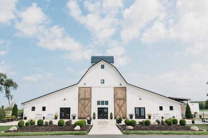 A large, white barn with a classic A-frame design, featuring dark accents and a large wooden door at the center. The barn is surrounded by well-manicured plants and shrubs in the foreground. A bright, clear blue sky with scattered clouds creates a vibrant backdrop, enhancing the barn's rustic yet modern aesthetic.