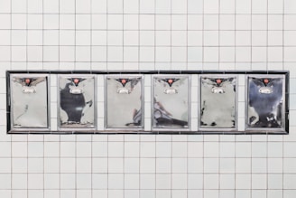 A series of six stainless steel mailbox doors are mounted on a tiled wall. Each mailbox has a small red emblem at the top and a silver handle below. The reflective surfaces of the mailboxes capture some abstract reflections.
