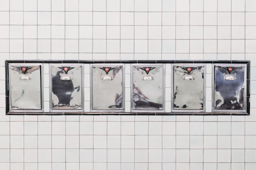 A series of six stainless steel mailbox doors are mounted on a tiled wall. Each mailbox has a small red emblem at the top and a silver handle below. The reflective surfaces of the mailboxes capture some abstract reflections.