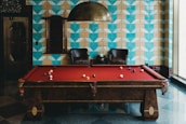 A stylish room features a red felt billiards table with balls scattered across its surface. The background includes a distinctive geometric blue and beige tiled wall, complemented by a metal ceiling lamp hanging overhead. Two dark leather chairs are situated against the wall, with small tables and a decorative item between them, creating an inviting, upscale atmosphere.