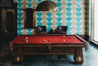 A stylish room features a red felt billiards table with balls scattered across its surface. The background includes a distinctive geometric blue and beige tiled wall, complemented by a metal ceiling lamp hanging overhead. Two dark leather chairs are situated against the wall, with small tables and a decorative item between them, creating an inviting, upscale atmosphere.