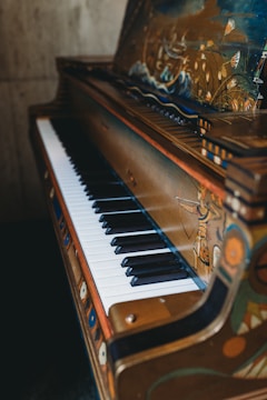 A piano with ornate, artistic designs on its exterior, including painted scenes and decorations. The piano keys alternate between white and black, and the frame has a rich, antique look. The artwork includes depictions of nature, human figures, and intricate patterns.