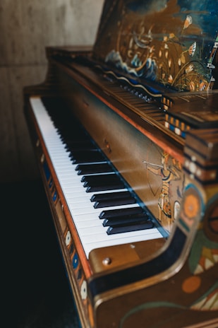 A piano with ornate, artistic designs on its exterior, including painted scenes and decorations. The piano keys alternate between white and black, and the frame has a rich, antique look. The artwork includes depictions of nature, human figures, and intricate patterns.