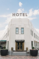 A large, elegant hotel building with an Art Deco architectural style, featuring a grand entrance labeled Bottleworks Hotel. There is a prominent sign on the rooftop spelling 'HOTEL' in large, illuminated letters. The structure is made of white tiles with decorative elements and large windows. Two black planters with green plants and colorful flowers are positioned symmetrically in front of the entrance.