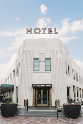 A large, elegant hotel building with an Art Deco architectural style, featuring a grand entrance labeled Bottleworks Hotel. There is a prominent sign on the rooftop spelling 'HOTEL' in large, illuminated letters. The structure is made of white tiles with decorative elements and large windows. Two black planters with green plants and colorful flowers are positioned symmetrically in front of the entrance.