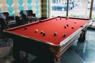 A billiards table with a red felt surface is set in a well-lit room with patterned walls and large windows. Several pool balls are scattered across the table. Two leather chairs sit adjacent to the table, and a glimpse of outside traffic can be seen through the windows.