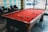 A billiards table with a red felt surface is set in a well-lit room with patterned walls and large windows. Several pool balls are scattered across the table. Two leather chairs sit adjacent to the table, and a glimpse of outside traffic can be seen through the windows.