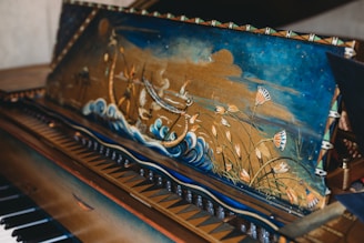 A beautifully decorated piano lid featuring a vibrant painting of a sailing ship on tumultuous waves, surrounded by floral elements and a sky with swirling clouds. The intricate artwork includes detailed flowers and dynamic elements of the sea.