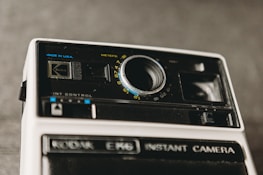 A vintage Kodak instant camera is prominently featured, showcasing its lens and various control knobs against a blurred background. The camera has a classic design with black and white elements, and the brand name is visibly printed.
