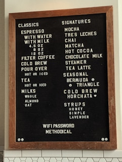 A stylish coffee shop menu board with a wooden frame features a variety of drink options. The left column lists 'Classics' including espresso with choices of water or milk, filter coffee, cold brew, pour-over, and tea. Various milk alternatives are also offered, such as whole, almond, and oat. The right column presents 'Signatures' like mocha, tres leches, chai, matcha, and hot cocoa, along with unique options such as Bermuda, Triangle, Cold Brew Horchata, and assorted syrups. The board also displays the WiFi password: Methodical.