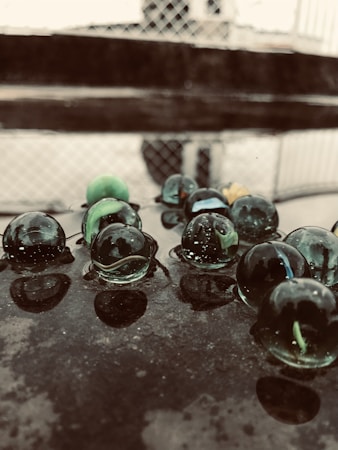 Glass marbles are scattered across a reflective, dark surface. The marbles are translucent with shades of green and blue, and they cast faint reflections on the surface. In the background, a blurred grid-like structure and horizontal lines are visible, likely part of a fence or railing.