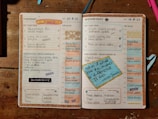 An open planner displaying a detailed schedule for Tuesday and Wednesday. The layout includes sections for top tasks, time blocks from 5 AM to 8 PM, other activities, and areas for reflection on successes and improvements. Decorative elements like motivational quotes and colorful stickers enhance the appearance. The dominant background includes a wooden surface and some writing utensils.