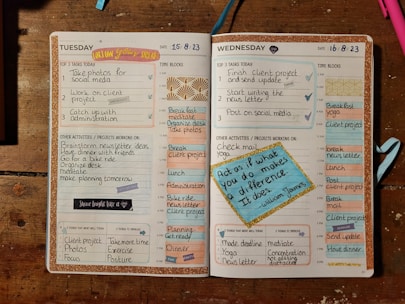 An open planner displaying a detailed schedule for Tuesday and Wednesday. The layout includes sections for top tasks, time blocks from 5 AM to 8 PM, other activities, and areas for reflection on successes and improvements. Decorative elements like motivational quotes and colorful stickers enhance the appearance. The dominant background includes a wooden surface and some writing utensils.