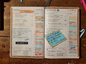 An open planner displaying a detailed schedule for Tuesday and Wednesday. The layout includes sections for top tasks, time blocks from 5 AM to 8 PM, other activities, and areas for reflection on successes and improvements. Decorative elements like motivational quotes and colorful stickers enhance the appearance. The dominant background includes a wooden surface and some writing utensils.