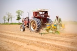 A farmer operates a red tractor equipped with a seed planting machine on a large field. The soil appears freshly plowed, with visible rows ready for planting. In the background, there are a few green trees against a clear blue sky.