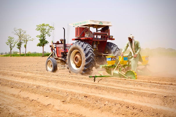 A farmer operates a red tractor equipped with a seed planting machine on a large field. The soil appears freshly plowed, with visible rows ready for planting. In the background, there are a few green trees against a clear blue sky.