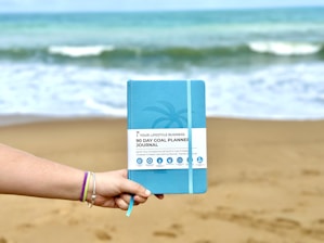A hand wearing colorful bracelets holds a blue 90-day goal planner journal against a sandy beach backdrop with waves in the ocean.