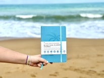 A hand wearing colorful bracelets holds a blue 90-day goal planner journal against a sandy beach backdrop with waves in the ocean.