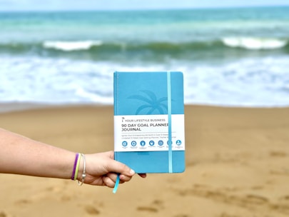 A hand wearing colorful bracelets holds a blue 90-day goal planner journal against a sandy beach backdrop with waves in the ocean.