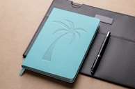 A turquoise notebook with an embossed palm tree design sits on a brown leather portfolio. A black pen with silver accents is placed alongside. The background is a neutral taupe surface.