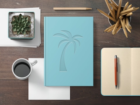 A neatly arranged desktop features a light blue notebook with an embossed palm tree design, placed centrally on a wooden surface. To the left, there is a white coffee cup filled with black coffee, accompanied by a green succulent plant in a square pot. On the right, an open blank notebook with an orange pen is visible, along with a container filled with sharpened pencils.