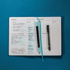 An open planner displaying a detailed weekly schedule with to-do lists on a blue surface. The left page is filled with handwritten tasks for Tuesday, including top three tasks and additional projects. The right page is Wednesday's schedule, mostly blank, with some text. Two pens, one black and one blue, are placed on top of the planner, and a blue ribbon bookmark is laying across the pages.