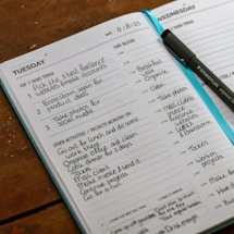 An open planner displaying a detailed schedule for Tuesday and Wednesday, including tasks such as creating freelance accounts and organizing activities. A pen is placed on the planner, and the pages are filled with handwritten notes outlining tasks, time blocks, and projects.