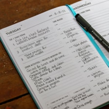 An open planner displaying a detailed schedule for Tuesday and Wednesday, including tasks such as creating freelance accounts and organizing activities. A pen is placed on the planner, and the pages are filled with handwritten notes outlining tasks, time blocks, and projects.