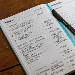 An open planner displaying a detailed schedule for Tuesday and Wednesday, including tasks such as creating freelance accounts and organizing activities. A pen is placed on the planner, and the pages are filled with handwritten notes outlining tasks, time blocks, and projects.