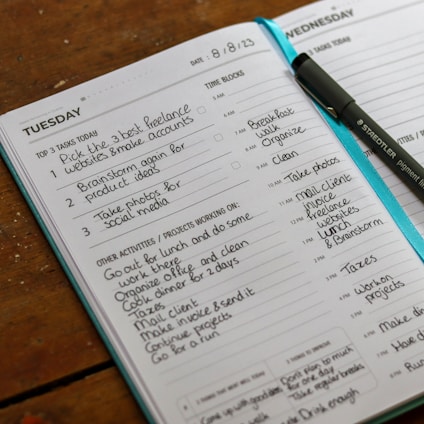 An open planner displaying a detailed schedule for Tuesday and Wednesday, including tasks such as creating freelance accounts and organizing activities. A pen is placed on the planner, and the pages are filled with handwritten notes outlining tasks, time blocks, and projects.