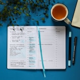 A two-page planner is open displaying a weekly schedule with handwritten tasks and appointments for Tuesday and Wednesday. It rests on a blue surface, accompanied by a blue ribbon bookmark, a black pen, a blue marker, a mug filled with tea, and a leafy plant in the upper left corner. An additional open notebook is partially visible to the right.