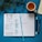 A two-page planner is open displaying a weekly schedule with handwritten tasks and appointments for Tuesday and Wednesday. It rests on a blue surface, accompanied by a blue ribbon bookmark, a black pen, a blue marker, a mug filled with tea, and a leafy plant in the upper left corner. An additional open notebook is partially visible to the right.